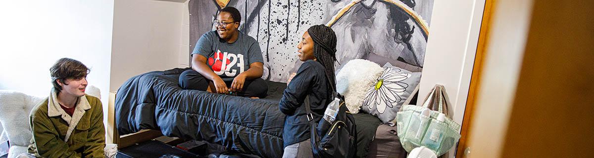 three students in residence hall; one on bed, one standing by bed, one sitting in folding chair